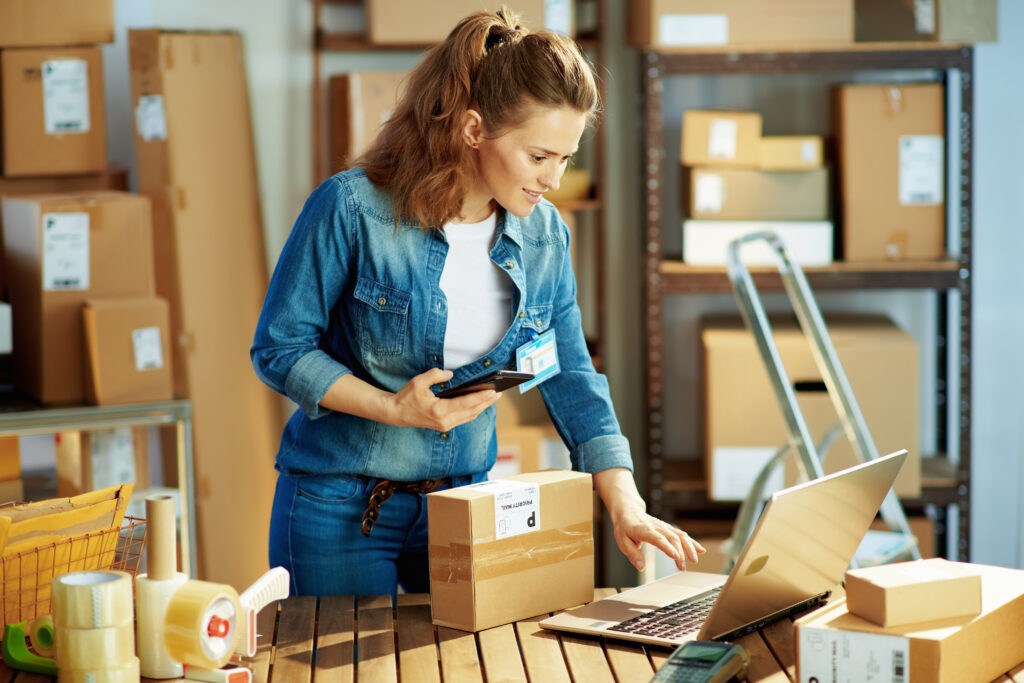 Woman in a warehouse looking at a laptop and holding a phone looking at a package