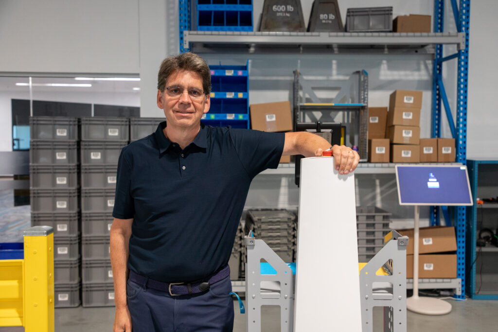 Person in a dark polo leaning on a LocusBot mast in a warehouse with shelving and totes behind