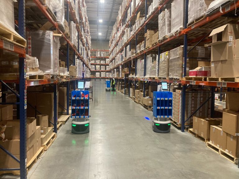 Two LocusBots carrying blue tote racks down a wide warehouse aisle of pallet racking, a worker in the distance