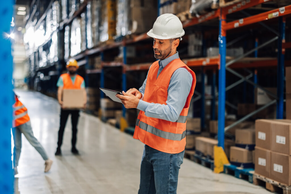 Warehouse supervisor in a hard hat and orange vest using a tablet as workers carry boxes