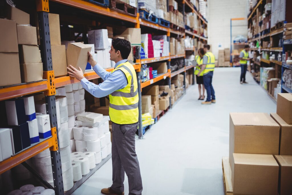 Worker in a safety vest stocking boxes on warehouse shelves while colleagues talk in the aisle