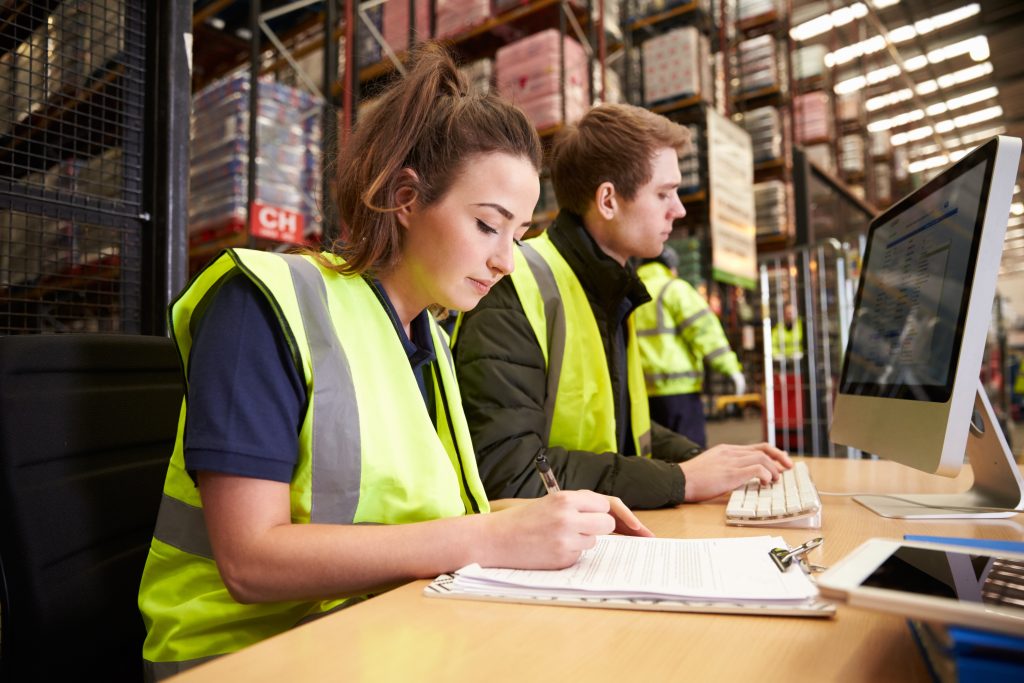 Warehouse staff in high-visibility vests doing paperwork and computer work at a desk inside a warehouse