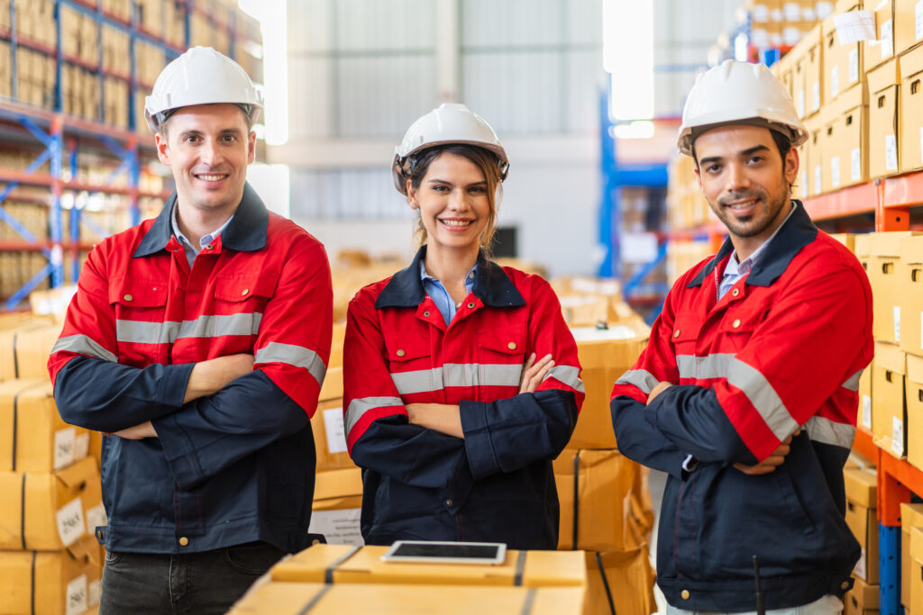 Three warehouse workers in hard hats and red safety uniforms standing with arms crossed