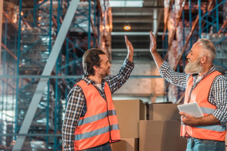 Two warehouse workers in orange safety vests giving a high five near stacked boxes