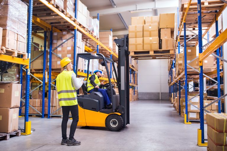 Warehouse worker with a clipboard directing a forklift operator between tall pallet racks