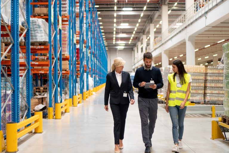 Three warehouse staff, one with a tablet, walking and talking down a wide aisle of tall pallet racks