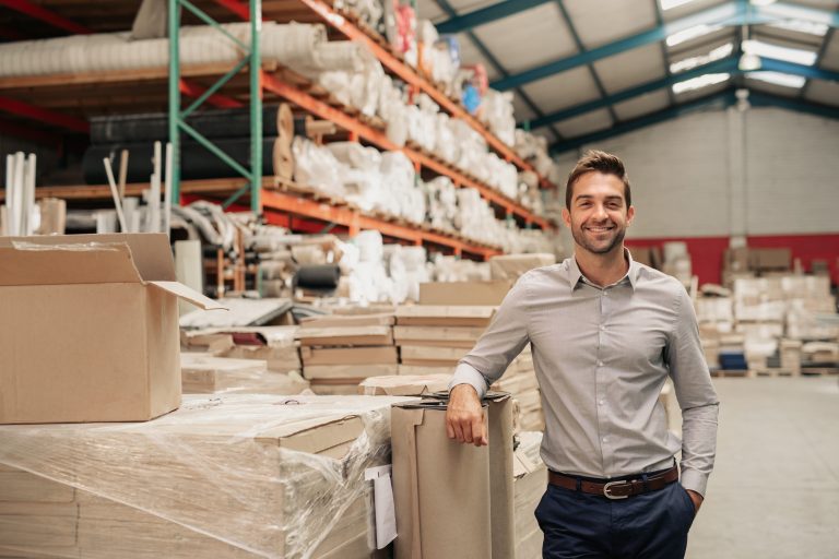 Smiling warehouse manager leaning on a box beside shelves stocked with wrapped goods