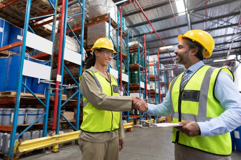 Two people in hard hats and safety vests shaking hands in a warehouse aisle
