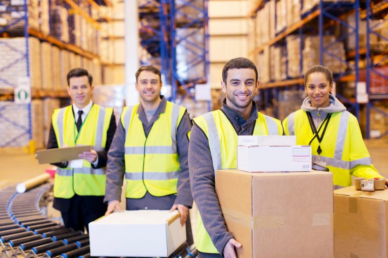 Four smiling warehouse workers in high-visibility vests holding boxes beside a roller conveyor