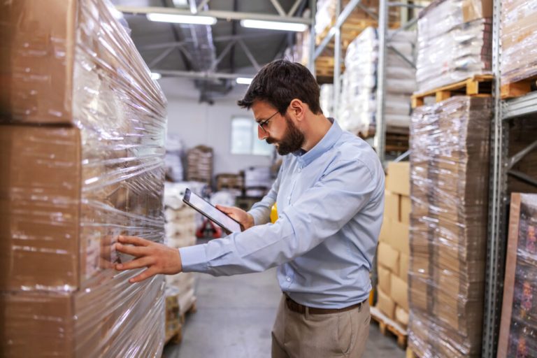 Worker checking shrink-wrapped pallets of boxes against a tablet in a warehouse
