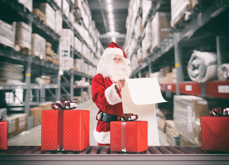 Santa Claus reading a long list beside red gift boxes on a conveyor in a warehouse aisle