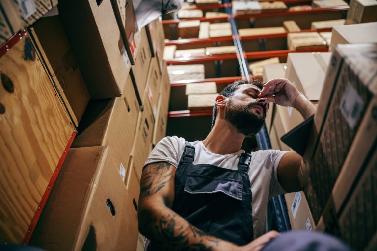 Tired warehouse worker in overalls rubbing their eyes while crouching between shelves of boxes