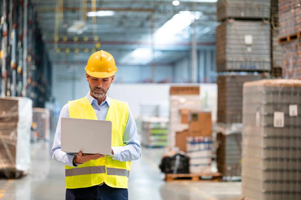 Warehouse manager in a hard hat and safety vest working on a laptop among shrink-wrapped pallets