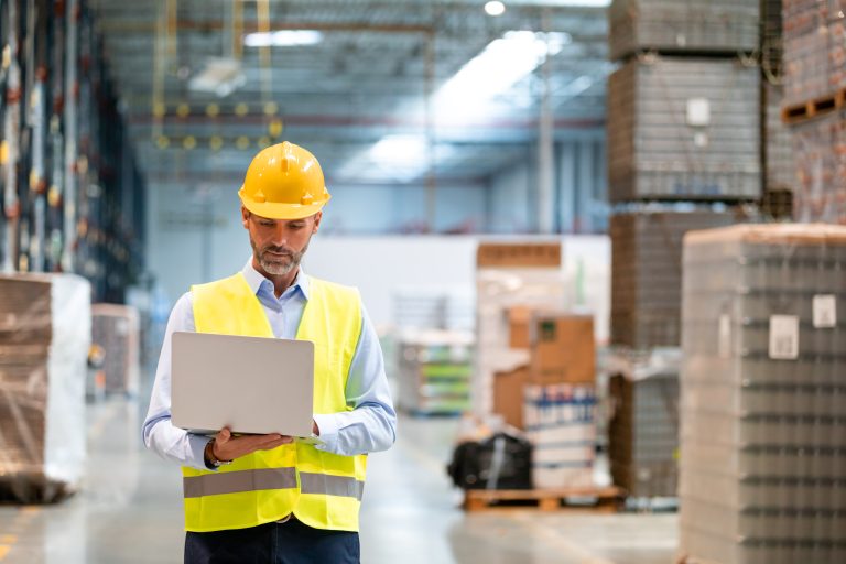 Warehouse manager in a hard hat and safety vest working on a laptop among shrink-wrapped pallets