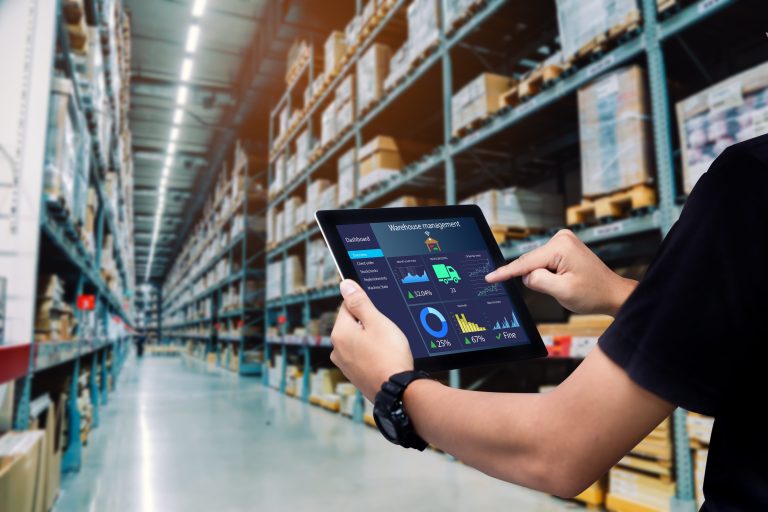 Hands holding a tablet showing a warehouse management dashboard in a long aisle of stocked shelving