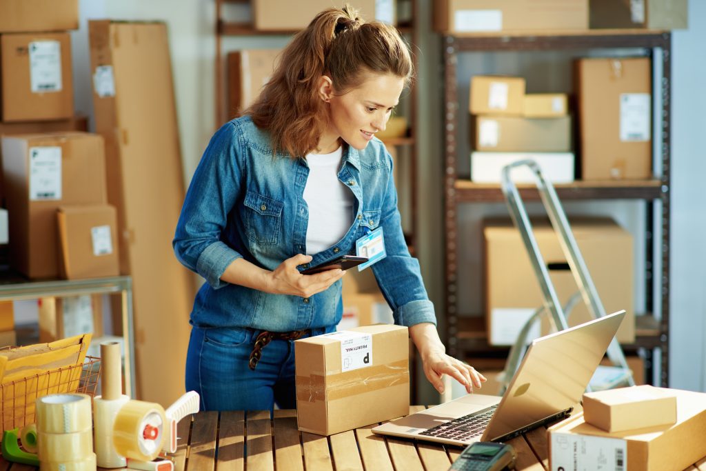 Small business owner holding a phone and typing on a laptop beside packed shipping boxes and tape