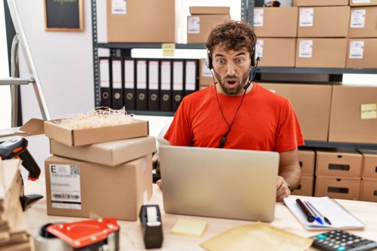 Surprised worker wearing a headset at a laptop in a small shipping office surrounded by boxes