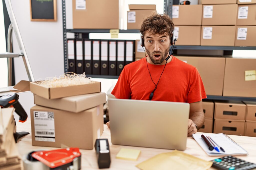 Surprised worker wearing a headset at a laptop in a small shipping office surrounded by boxes
