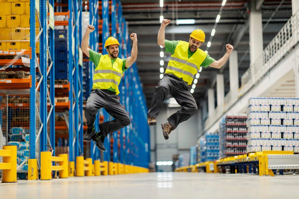 Two cheering warehouse workers in hard hats and safety vests jumping in the air in a racked aisle