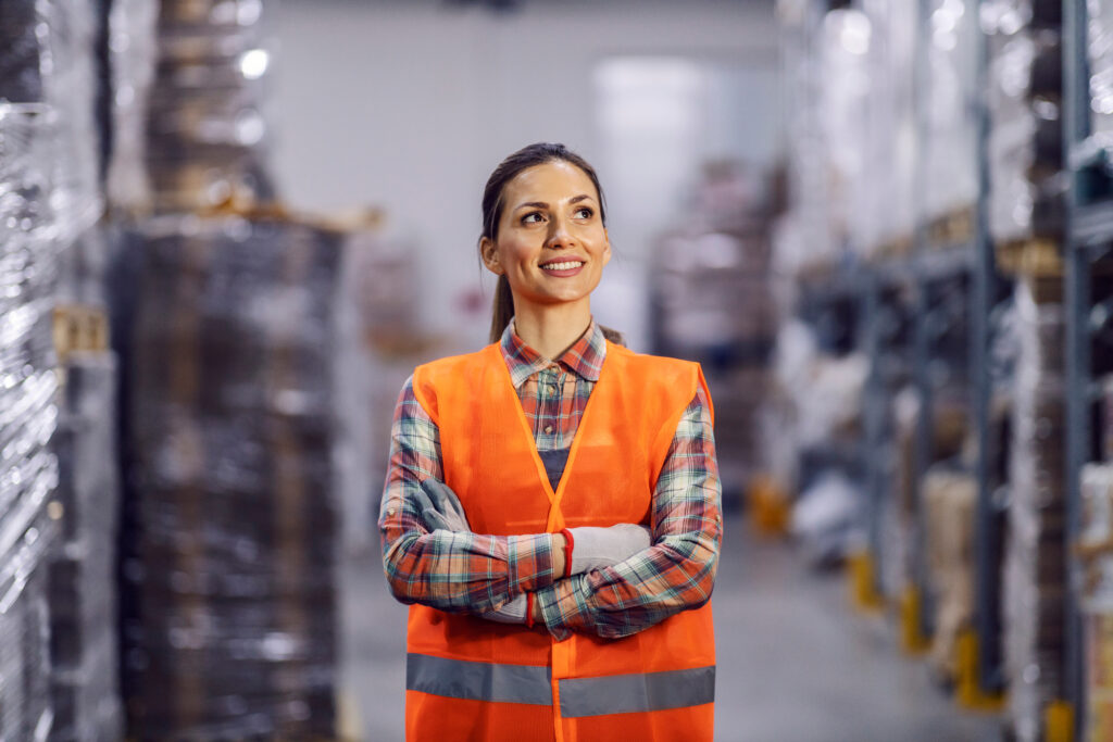 Smiling warehouse worker in an orange safety vest standing with arms crossed between storage racks