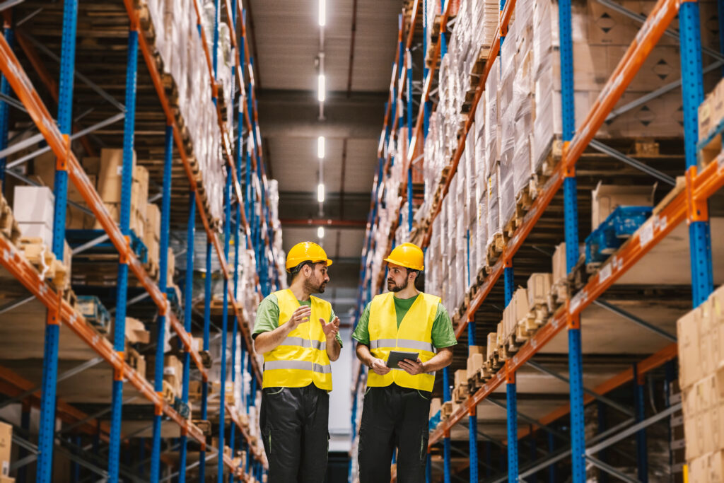 Two workers in hard hats and safety vests talking with a tablet between tall pallet racks