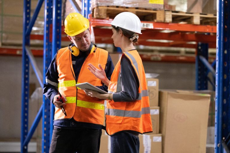 Two warehouse workers in hard hats and orange safety vests discussing plans on a tablet between racks