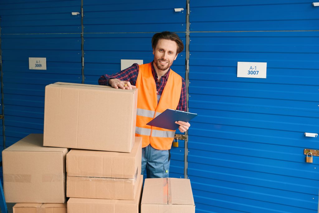 Smiling worker in an orange safety vest with a clipboard beside stacked boxes and blue storage units
