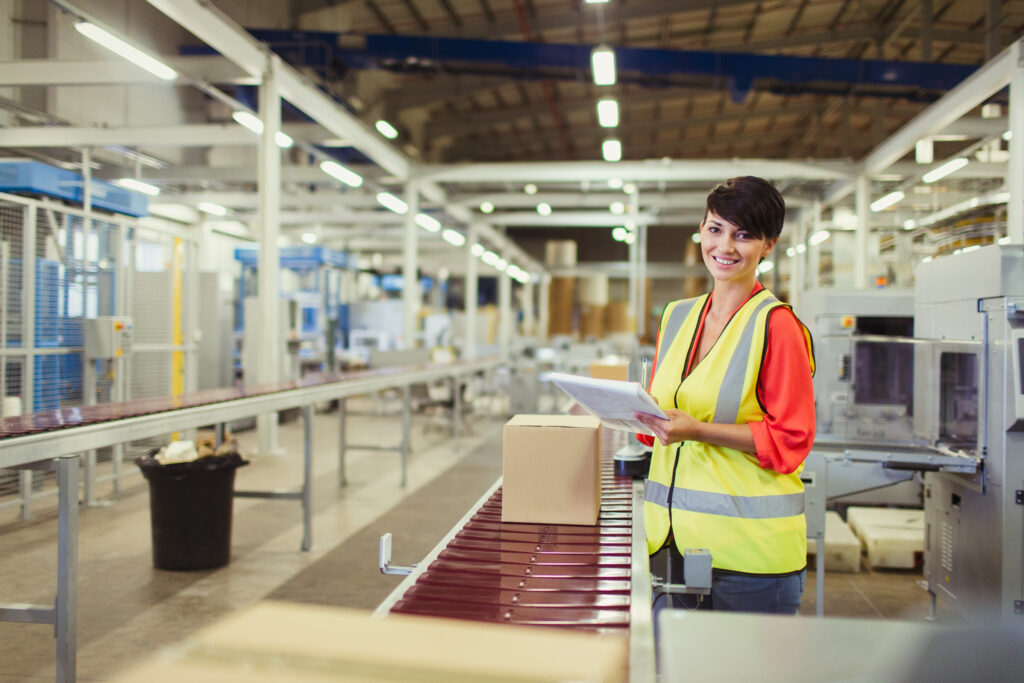 Smiling warehouse worker in a high-visibility vest holding a clipboard beside a box on a conveyor