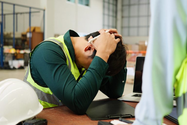 Stressed warehouse supervisor in a safety vest holding their head at a desk with a laptop and hard hat