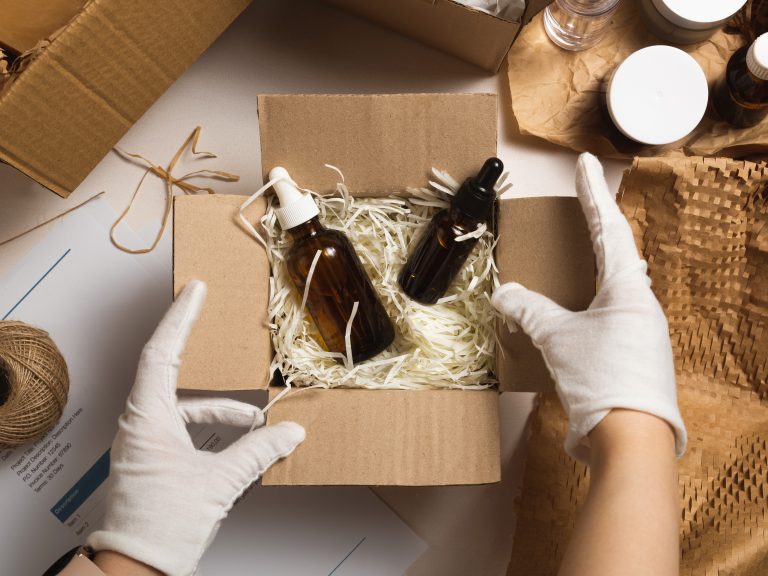 Gloved hands packing amber dropper bottles into a cardboard box filled with paper shred