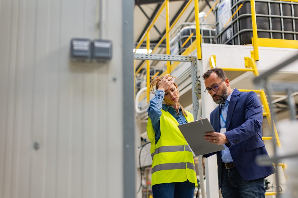 Stressed warehouse worker in a safety vest holding her head while a manager reviews a clipboard