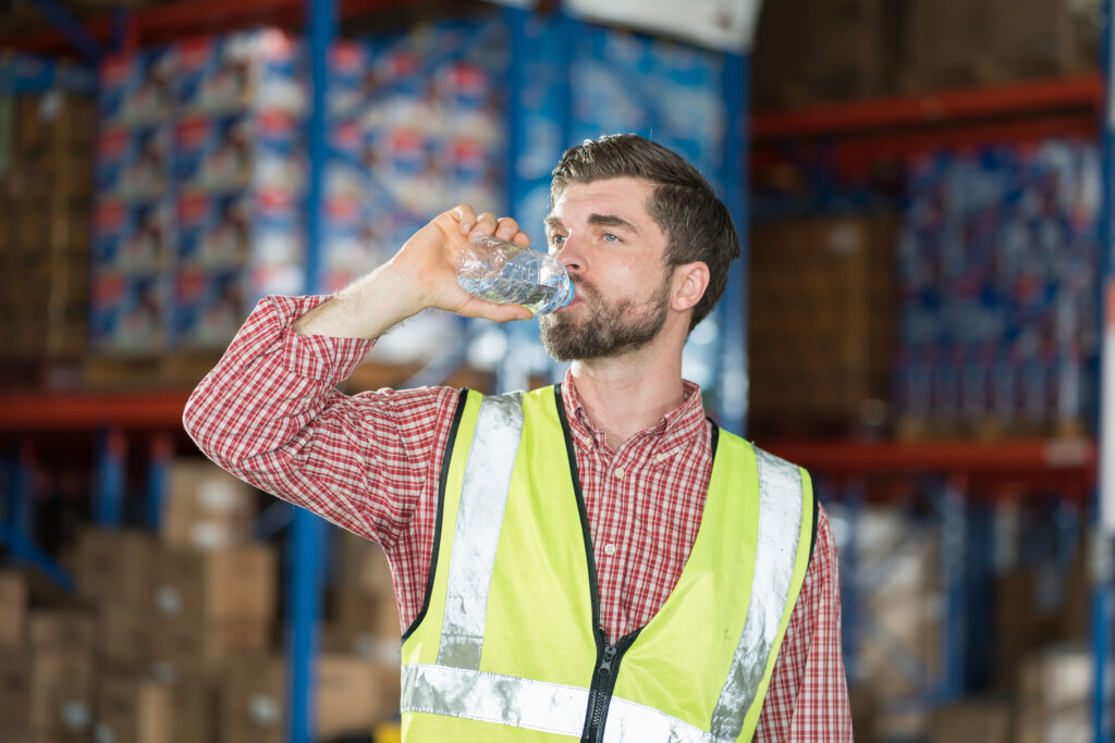 Warehouse worker in a high-visibility vest drinking from a water bottle in front of stocked shelving