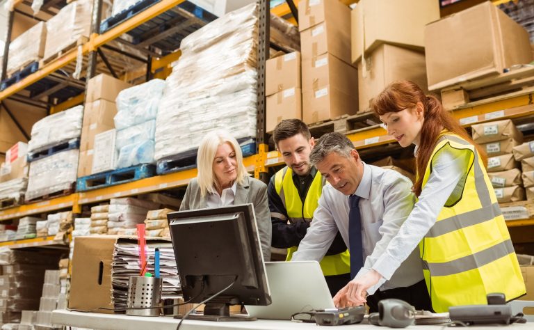 Warehouse team gathered around a laptop and computer at a workstation among stocked shelves
