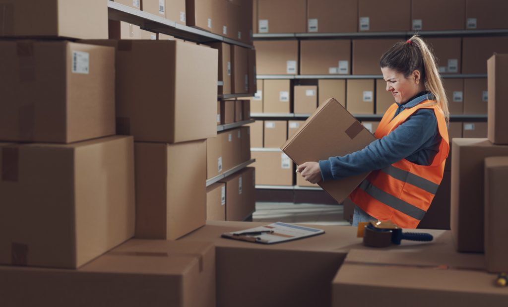 Frustrated warehouse worker in an orange safety vest lifting a cardboard box among stacked parcels
