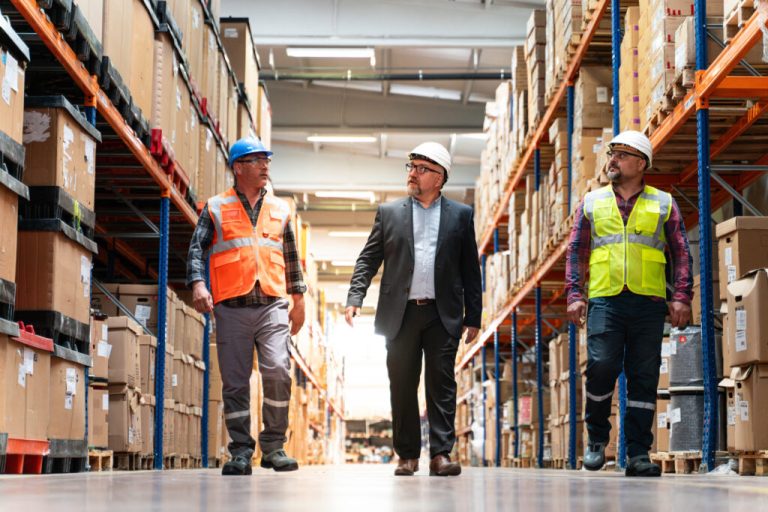 Manager in a suit walking a warehouse aisle with two workers in hard hats and safety vests