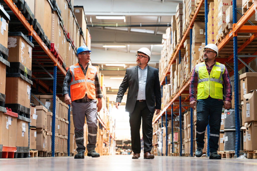 Manager in a suit walking a warehouse aisle with two workers in hard hats and safety vests