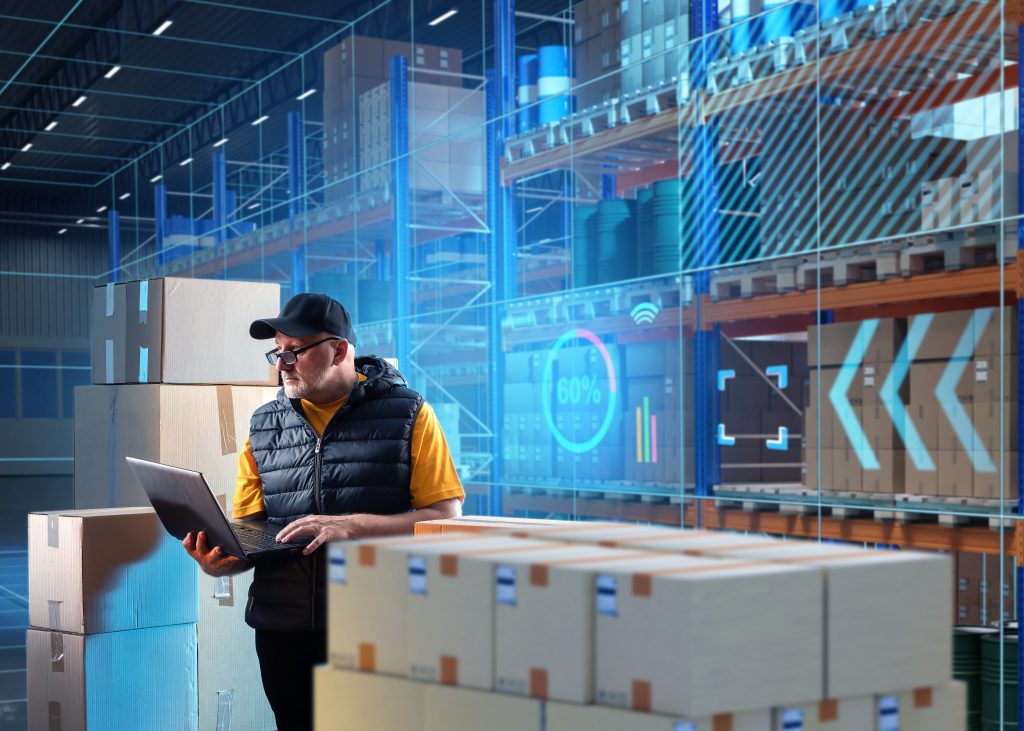 Warehouse worker in a cap using a laptop among stacked boxes, with digital inventory data overlays on the shelving