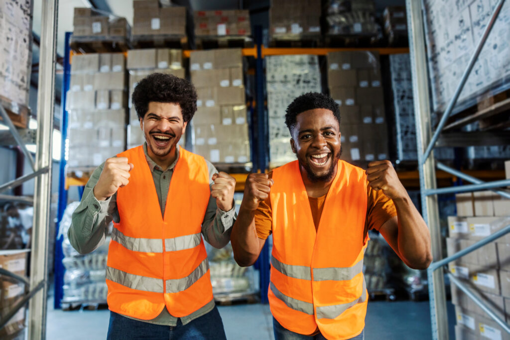 Two cheering warehouse workers in orange safety vests celebrating in a storage aisle