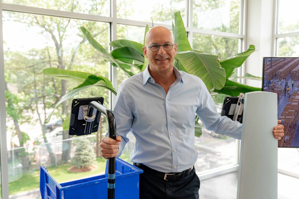 Al Dekin smiling with hands on two LocusBots, one carrying a blue tote, in a bright office with large plants