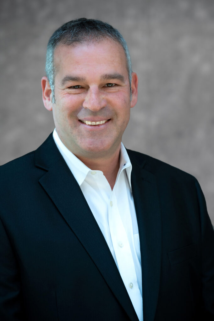 Headshot of Amit Levy, smiling in a dark suit jacket and white open-collar shirt against a gray backdrop