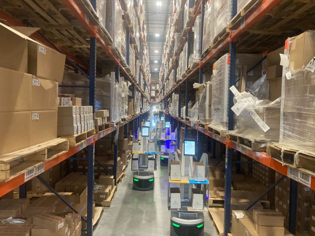 Several LocusBots with tote shelves and touchscreens moving down a narrow aisle of tall pallet racking at KSP