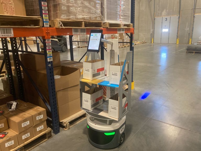 LocusBot carrying cardboard order boxes on a two-tier shelf beside pallet racking in a warehouse