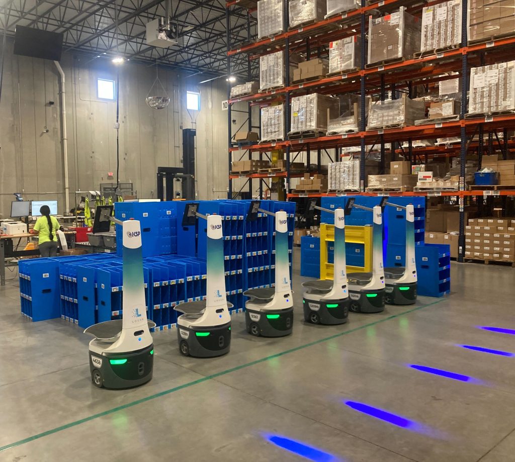 Row of KSP-branded LocusBots parked beside stacked blue totes in front of orange warehouse pallet racking