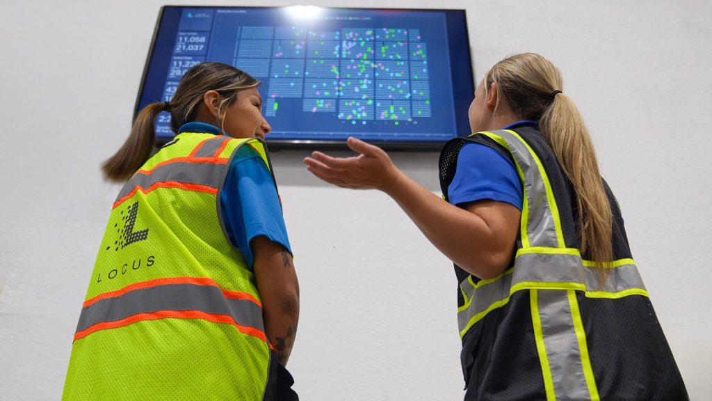Two warehouse associates in safety vests reviewing a Locus dashboard displayed on a wall screen