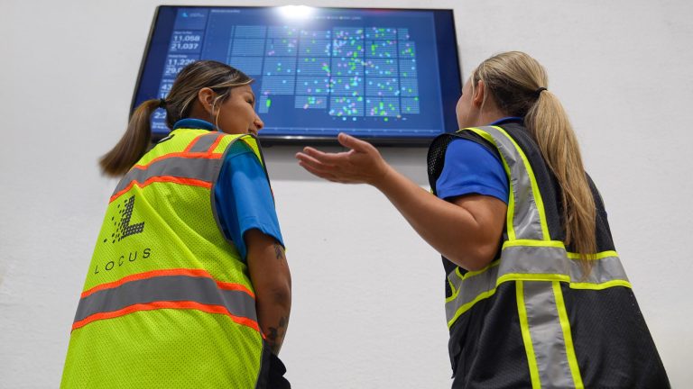 Two warehouse associates in safety vests reviewing a Locus dashboard displayed on a wall screen