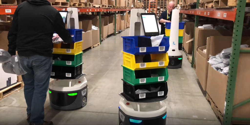 Workers picking items into colorful stacked totes carried by LocusBots in a warehouse aisle