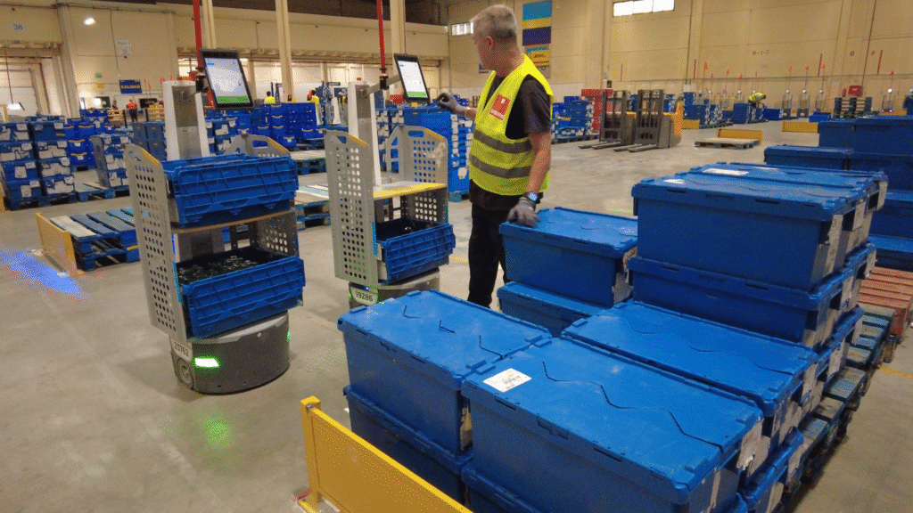 Worker in a high-visibility vest beside LocusBots carrying blue totes among stacks of blue crates