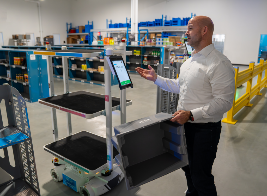 Person gesturing toward a Locus Vector robot with shelf platforms and touchscreen in a demo warehouse