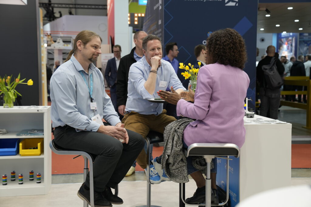 Three people seated at a high table talking at a busy trade show booth decorated with yellow tulips
