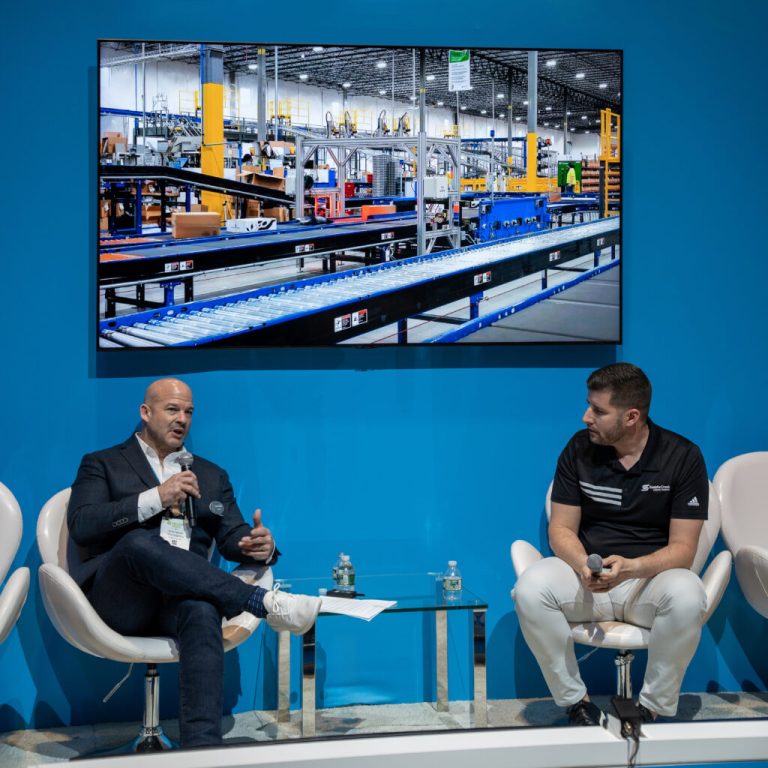 Two speakers with microphones in a panel discussion on a blue stage, a warehouse conveyor photo on screen behind